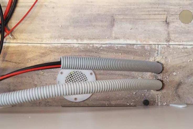 Zwei graue Wellschläuche über einer runden weißen Abdeckung mit Lochmuster auf staubigem hellbraunem Untergrund; oben links rote und schwarze Kabel sichtbar; rechts ein rundes Bohrloch; unten rechts weiße Oberfläche