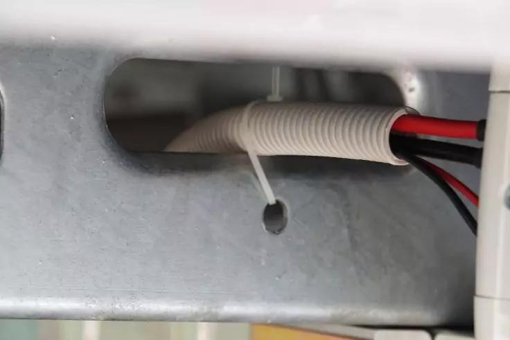 Weißer Wellschlauch mit roten und schwarzen Kabeln, mit einem weißen Kabelbinder zusammengebunden, verläuft durch eine rechteckige Öffnung in einer Metallplatte; darunter ein kleines rundes Loch