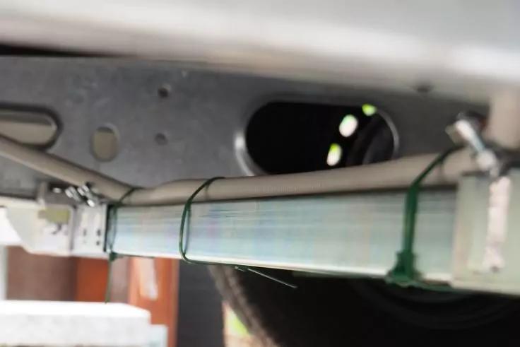 Blick auf den Unterboden mit einem horizontal an einem Metallträger befestigten Metallrohr, grünen Kabelbindern, einem Reifen und dem Boden im Hintergrund