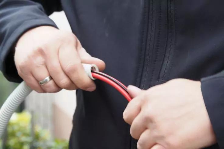 Zwei Hände halten einen grauen Wellschlauch und zwei freiliegene Kabel (rot und schwarz); Person trägt eine dunkle Jacke