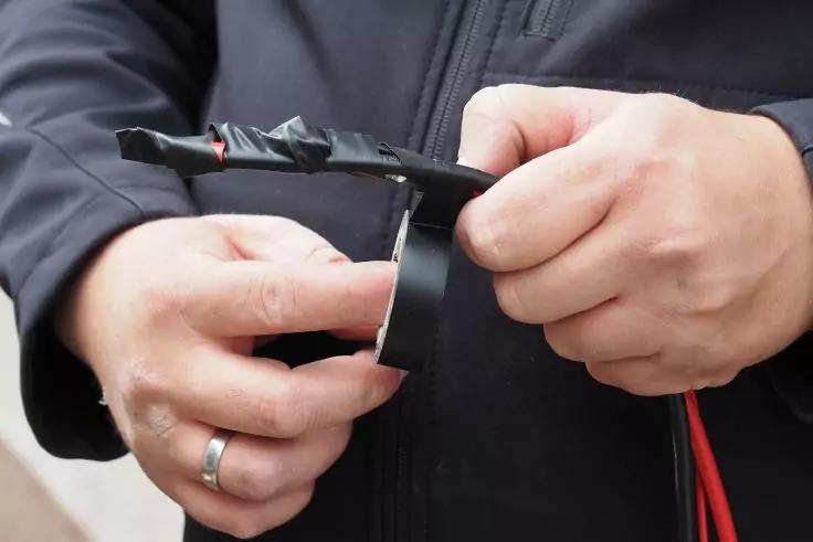 Zwei Hände halten ein schwarzes Kunststoffteil mit roten und schwarzen Kabeln, die mit schwarzem Klebeband umwickelt sind; linke Hand mit Ring, Person trägt eine dunkle Jacke