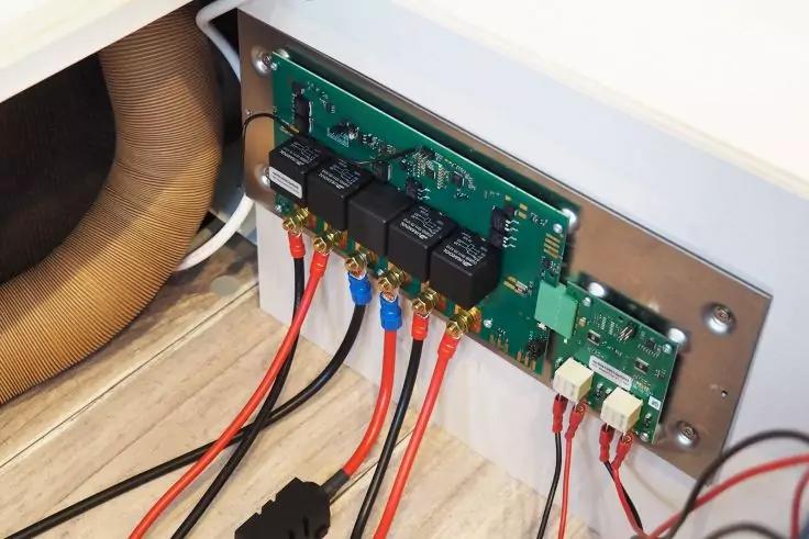 Grüne Leiterplatte mit mehreren schwarzen Relais und weißen Steckern auf einer Metallplatte; mehrere rote und schwarze Kabel mit Ringkabelschuhen angeschlossen; beige Schlauch und weißer Möbelrand im Hintergrund