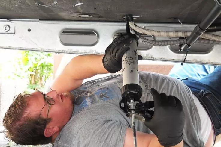 Person liegt auf dem Rücken unter einem Fahrzeug und hält mit beiden Händen ein Rohr; trägt schwarze Handschuhe, Brille und graues T‑Shirt; Fahrzeugunterseite und Kabel sichtbar