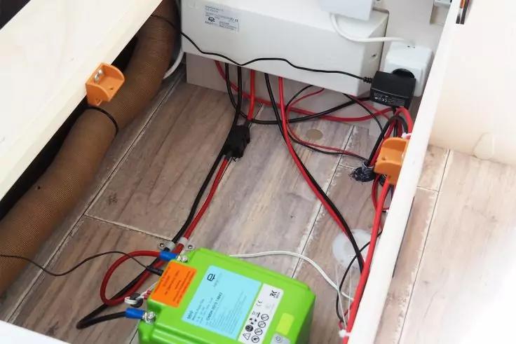 Offenes Bodenfach mit grünem rechteckigem Gerät mit Aufklebern, mehreren roten und schwarzen Kabeln, weißem Elektronikgehäuse an der Rückwand, orangen Steckverbindern, braunem Rohr und hellem Holzfußboden