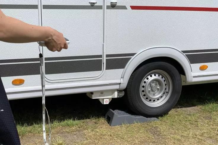 Teilansicht eines weißen Wohnwagens mit grauen und roten Streifen, sichtbares Rad mit schwarzem Unterlegkeil unter dem Reifen; links eine Hand hält einen weißen Gurt, Grasboden sichtbar