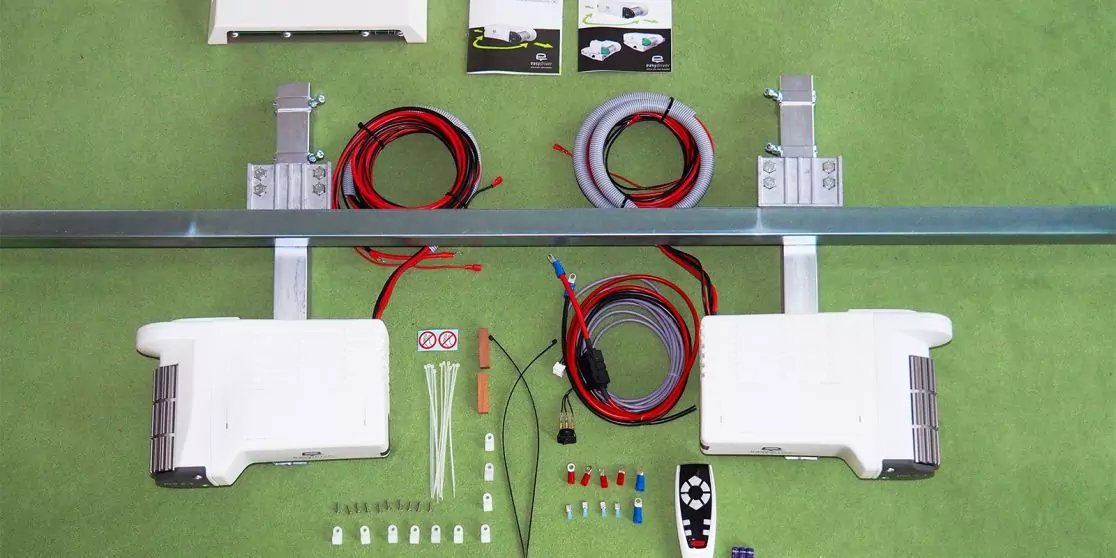 Zwei wei&szlig;e rechteckige Ger&auml;te, Metall-Montageschienen mit Halterungen, mehrere aufgerollte rote und schwarze Kabel, verschiedene kleine Steckverbinder, Schrauben und Kabelschuhe, zwei kleine Karten oben und ein kleines schwarz-wei&szlig;es Bedienger&auml;t rech