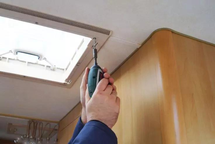 Zwei H&auml;nde halten einen gr&uuml;nen Akkuschrauber an den Rahmen einer ge&ouml;ffneten Dachluke in einer Innenraumdecke, rechts ein helles Holzpaneel