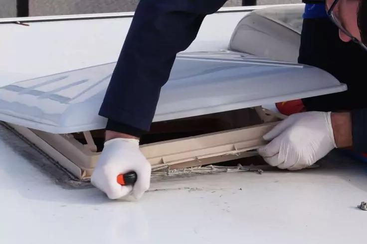 Wei&szlig;er Kunststoff-Dachlukenabdeckung, halb ge&ouml;ffnet auf einer wei&szlig;en Dachfl&auml;che; zwei H&auml;nde mit wei&szlig;en Handschuhen halten einen Schraubendreher mit orange‑schwarzem Griff; dunkle Jacken&auml;rmel und Teil einer Person sichtbar