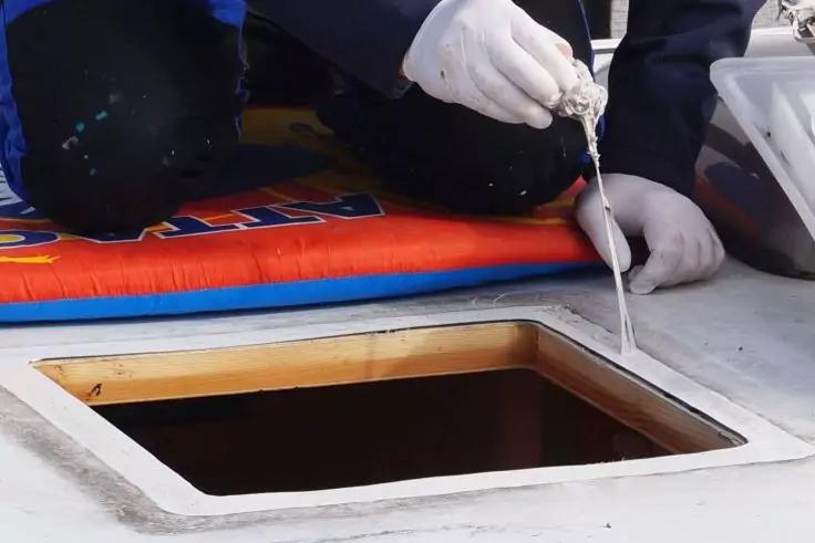 Zwei H&auml;nde in wei&szlig;en Handschuhen halten eine Kartuschenpistole und dr&uuml;cken wei&szlig;es Material an die Kante einer rechteckigen Luke in einer wei&szlig;en Deckfl&auml;che; rechts eine wei&szlig;e Kunststoffbox, im Hintergrund eine bunte Sitzauflage