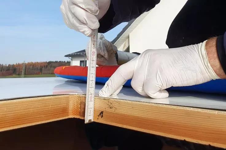 Zwei H&auml;nde mit wei&szlig;en Handschuhen halten ein senkrechtes Metallma&szlig;band an der Kante einer Holzplatte; auf der Oberfl&auml;che liegen ein rotes und ein blaues Band, im Hintergrund ein Geb&auml;ude und B&auml;ume