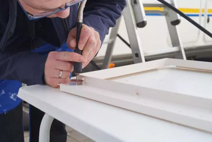 Person h&auml;lt ein schwarzes Handwerkzeug mit Metallspitze &uuml;ber einem wei&szlig;en rechteckigen Rahmen auf einem Tisch; Teil des Oberk&ouml;rpers mit dunkelblauer Jacke und Brille sowie Leiter und Kabel im Hintergrund sichtbar