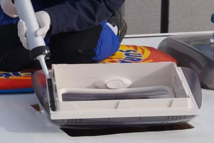 Hand mit wei&szlig;em Handschuh h&auml;lt wei&szlig;en Schlauch, der an einem rechteckigen wei&szlig;en Kunststoffaufsatz mit runder Vertiefung befestigt ist; im Hintergrund eine Person mit dunkelblauer Jacke und blauer Hose, bunte Unterlage und wei&szlig;e Fl&auml;che sichtbar