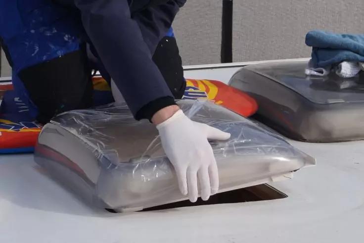 Zwei H&auml;nde mit wei&szlig;en Handschuhen pressen eine durchsichtige, mit Folie bedeckte rechteckige Kuppel &uuml;ber eine rechteckige &Ouml;ffnung in einer wei&szlig;en Dachfl&auml;che; im Hintergrund eine zweite &auml;hnliche Kuppel, ein blauer Lappen und eine knieende Person in du