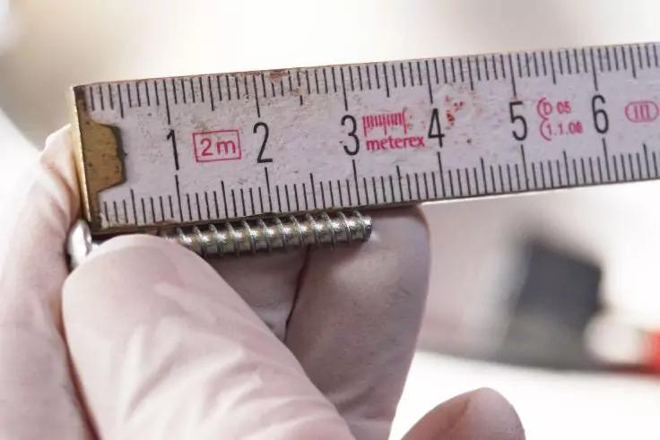 Hand in wei&szlig;em Handschuh h&auml;lt Metallschraube neben Ma&szlig;band mit Zentimeter-Skala