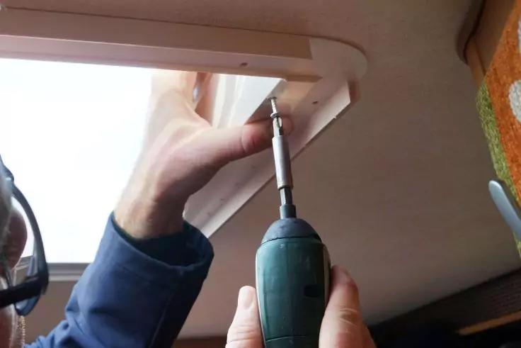 Hand h&auml;lt gr&uuml;nen Akkuschrauber und setzt Schraube in wei&szlig;en Rahmen einer rechteckigen Dachluke; zweite Hand st&uuml;tzt Rahmen; Teil eines Gesichts mit Brille links, orange gemustertes Polster rechts sichtbar