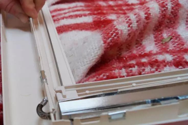 Hand h&auml;lt eine schmale wei&szlig;e Stange neben einem wei&szlig;en Rahmen mit Metallbeschl&auml;gen; rot-wei&szlig; gemustertes Stricktuch im Hintergrund; kleine schwarze Spiralfeder am Rahmen