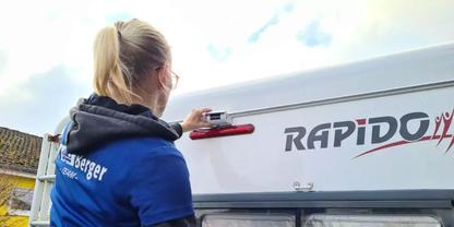 Person mit blondem Pferdeschwanz, von hinten, mit Brille, dunkler Kapuzenjacke unter blauem Shirt, ber&uuml;hrt Griff neben rotem R&uuml;cklicht an wei&szlig;er Heckklappe mit Schrift 'RAPIDO'