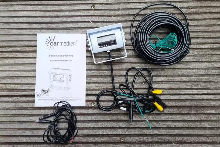 Bedienungsanleitung mit Schriftzug "carmedian", kleines rechteckiges Ger&auml;t mit St&auml;nder und angeschlossenem Kabel, mehrere aufgewickelte Kabelstr&auml;nge mit gr&uuml;nen und gelben Steckern, alles auf Holzlattenuntergrund