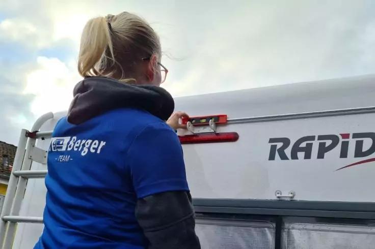 Person mit blondem Pferdeschwanz und Brille, tr&auml;gt blaues Shirt mit wei&szlig;em "Berger"-Logo, steht mit Hand an rotem Verschluss an einem wei&szlig;en Wohnwagen mit Schriftzug "RAPID", bew&ouml;lkter Himmel im Hintergrund