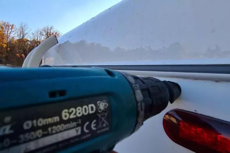 Nahaufnahme eines blauen Akkuschraubers mit Bohrspitze nahe an einer wei&szlig;en, gew&ouml;lbten Oberfl&auml;che; rechts ein rotes R&uuml;cklicht, im Hintergrund B&auml;ume und Himmel