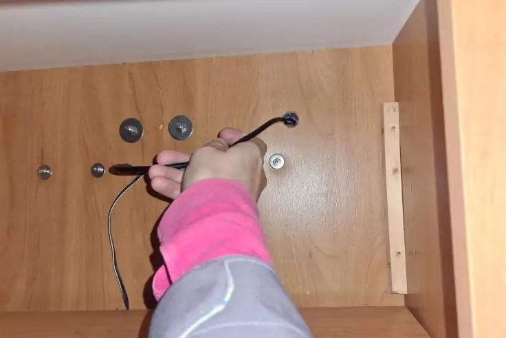 Innen eines hellbraunen Holzschranks, eine Hand in rosa-grauem &Auml;rmel h&auml;lt einen Schraubendreher an einer Metallbefestigung, mehrere runde Metall&ouml;ffnungen an der R&uuml;ckwand und ein schwarzes Kabel