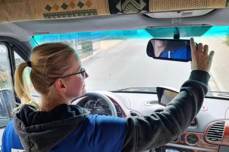 Person mit blondem Pferdeschwanz und Brille sitzt am Steuer eines Autos und stellt den R&uuml;ckspiegel ein; Hand ber&uuml;hrt den Spiegel, Armaturenbrett und Stra&szlig;e durch Windschutzscheibe sichtbar