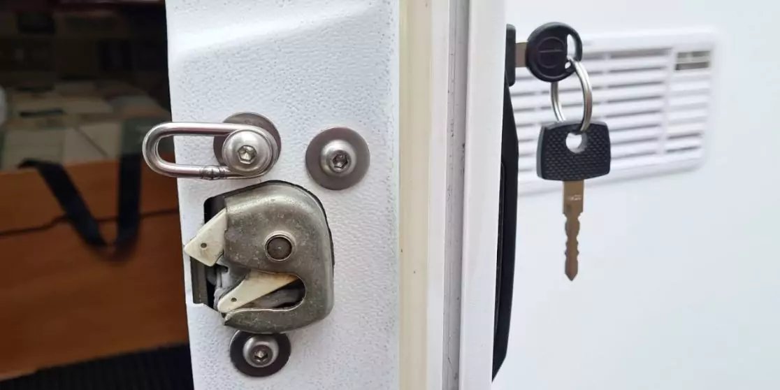 Wei&szlig;e T&uuml;rkante mit Metallriegel und Schlie&szlig;mechanismus links, rechts ein Schloss mit eingestecktem Schl&uuml;ssel und schwarzem Schl&uuml;sselanh&auml;nger an einer wei&szlig;en Platte, L&uuml;ftungsgitter rechts, Holzinnenraum im Hintergrund links