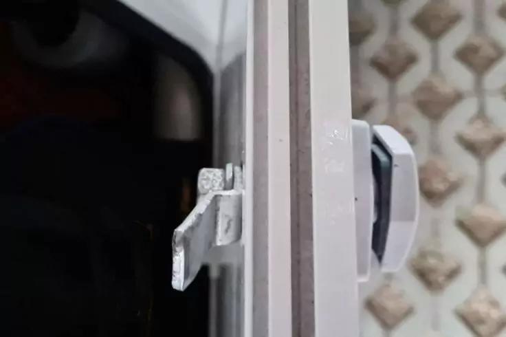 Nahaufnahme eines wei&szlig;en Kunststoffgriffs und eines metallischen Riegels an einem wei&szlig;en Fensterrahmen, rechts gemusterte Bodenfliesen, links dunkler Innenraum