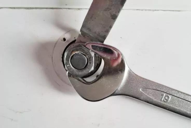 Silberner Ringschl&uuml;ssel mit Markierung '19' auf einer Mutter und Unterlegscheibe in einer runden Vertiefung auf wei&szlig;er Fl&auml;che; ein flaches Metallblech liegt am Bolzen