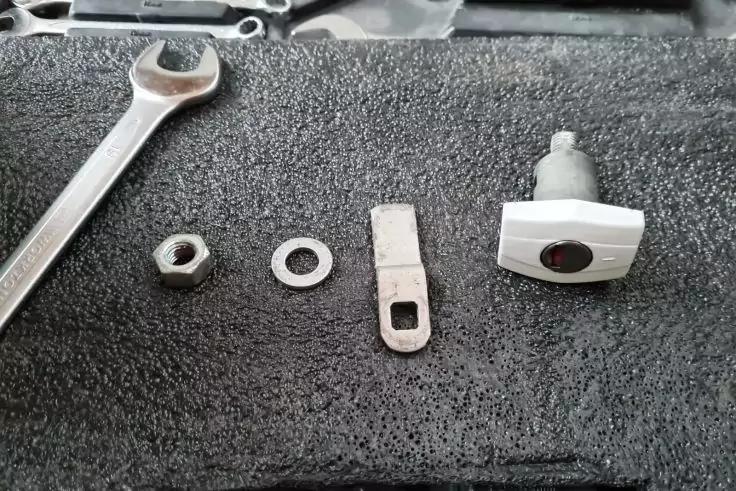 Auf schwarzer Unterlage: Maulschl&uuml;ssel, Sechskantmutter, Unterlegscheibe, flaches Metallst&uuml;ck mit rechteckiger &Ouml;ffnung und grauer Kunststoffknopf mit kurzem Gewindest&uuml;ck; oben am Rand ein Teil eines Werkzeugs