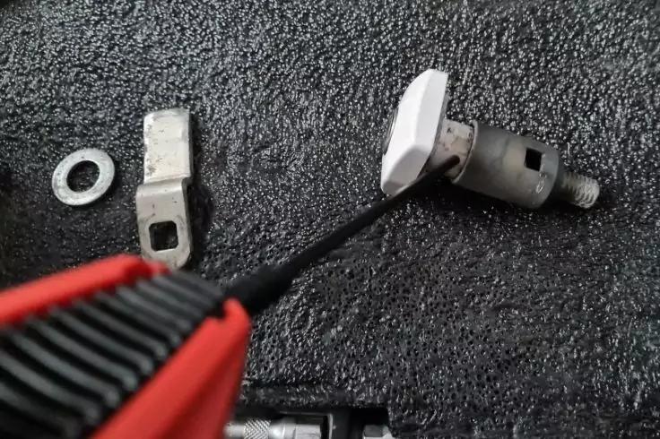 Zylinderf&ouml;rmiges Metallteil mit Schraube und wei&szlig;em Kunststoffst&uuml;ck, ein schwarzer Stab eines rot-schwarzen Werkzeugs ist darin eingesetzt; daneben eine Metallunterlegscheibe und ein gebogenes Metallst&uuml;ck auf einer schwarzen geriffelten Matte