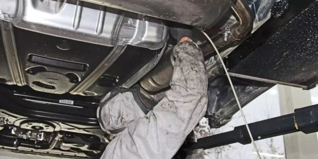 Person in grauer Arbeitskleidung unter einem angehobenen Auto, Arme nach oben, sichtbare Auspuffanlage und Fahrzeugunterboden