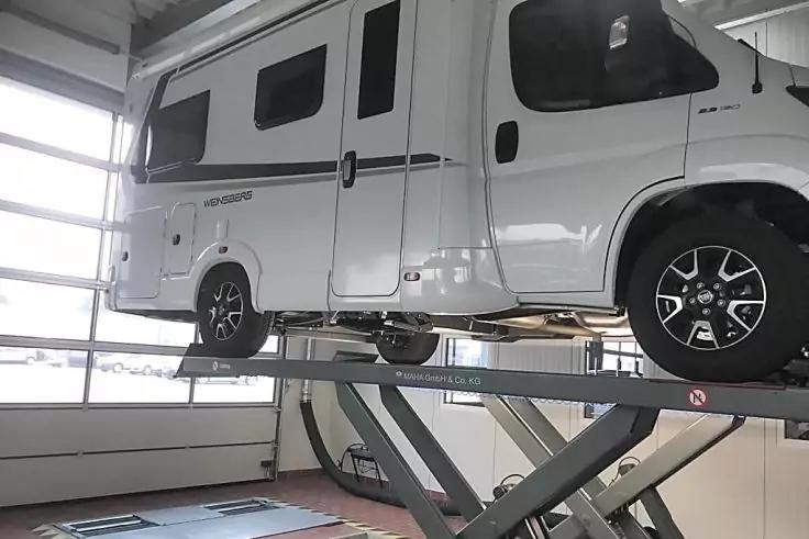 Wei&szlig;es Wohnmobil auf einer Scherenhebeb&uuml;hne in einer Werkstatt, sichtbare Seitenfenster und gro&szlig;e verglaste Garagentore im Hintergrund