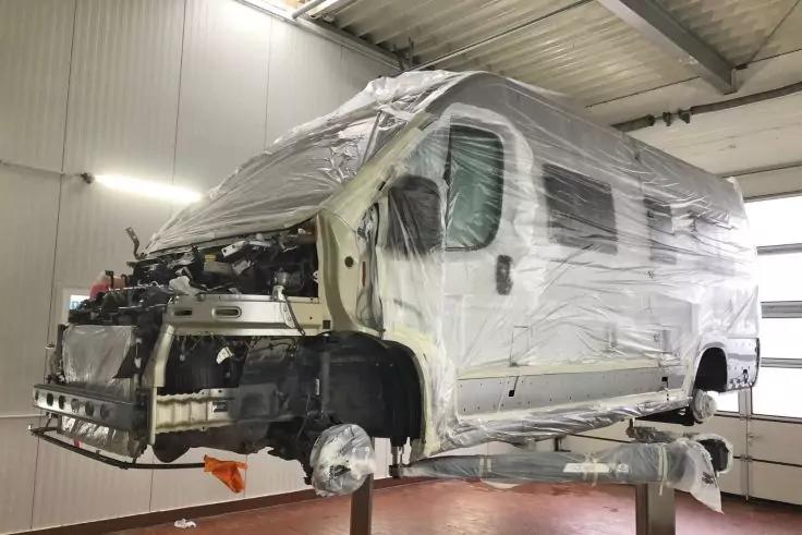 Wei&szlig;es Wohnmobil in einer Werkstatt auf Hebeb&uuml;hnen aufgehoben, vorderer Sto&szlig;f&auml;nger entfernt, Vorderachse ohne Rad sichtbar, Fahrzeug teilweise mit transparenter Folie abgedeckt