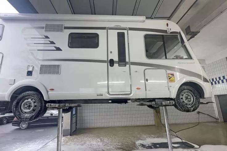 Wei&szlig;es Wohnmobil seitlich auf einer Hebeb&uuml;hne in einer Werkstatt, mit T&uuml;r, Fenstern und zwei sichtbaren R&auml;dern; geflieste W&auml;nde im Hintergrund