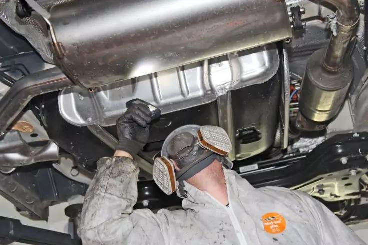 Person in wei&szlig;em Schutzanzug mit Handschuhen und Atemschutzmaske unter einem Fahrzeug; Auspuff und Fahrwerk sichtbar