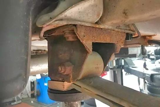 Unterboden eines Fahrzeugs, Nahaufnahme einer Blattfeder mit verschraubener Halterung und sichtbarem Rost, Metallbolzen, blauer Beh&auml;lter im Hintergrund