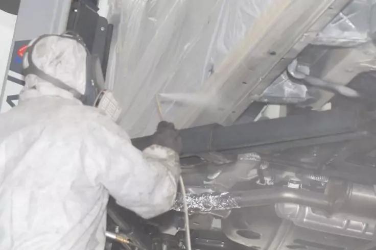 Person in wei&szlig;em Schutzanzug und Maske h&auml;lt eine Spritzpistole an der Unterseite eines Fahrzeugs