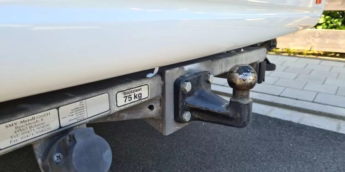 Nahaufnahme einer Anh&auml;ngerkupplung mit Kugel, elektrischer Steckdose und Aufkleber "75 kg" unter der wei&szlig;en Sto&szlig;stange