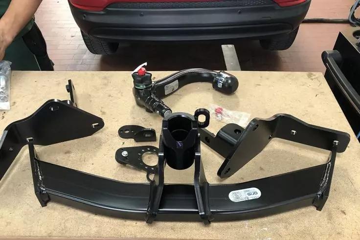 Mehrere schwarz lackierte Metallteile und eine runde Metallkugel auf einer Werkbank, kleine Plastiktüte mit Schrauben, linke Hand am Bildrand, Stoßstange eines roten Autos im Hintergrund