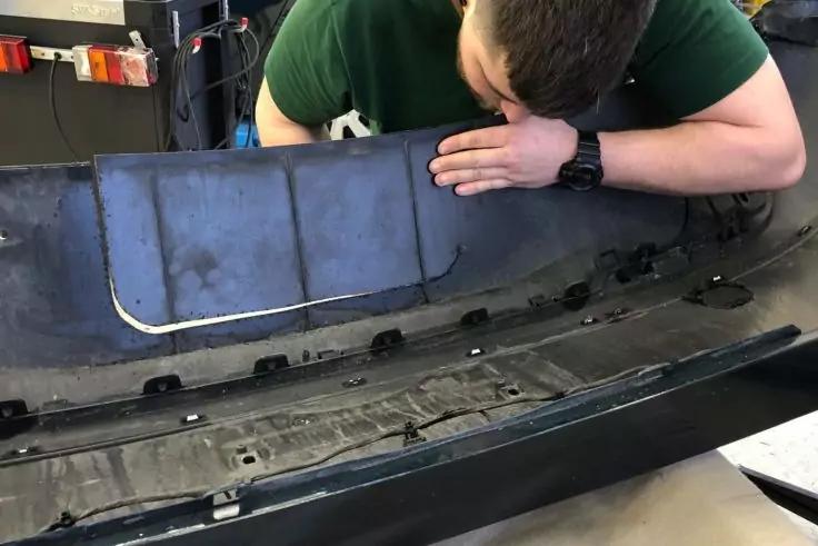 Person beugt sich über eine abgenommene schwarze Autostoßstange, Innenfläche mit Halterungen sichtbar; Armbanduhr am Handgelenk; Werkstatt im Hintergrund