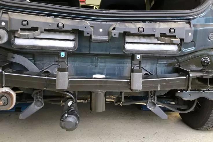 Heckansicht eines Fahrzeugs mit freiliegenden Metallträgern und Halterungen, zwei rechteckigen Leuchtelementen oberhalb, mittig angebrachte Anhängerkupplung, sichtbare Kabel, teilweise sichtbares rechtes Hinterrad, grauer Boden