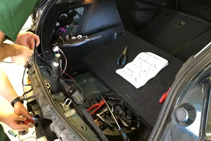 Offener Kofferraum eines Autos mit freigelegten Kabeln und Steckern, verschiedenen Werkzeugen und einem gefalteten Anleitungsblatt, links Hände mit grünem Ärmel