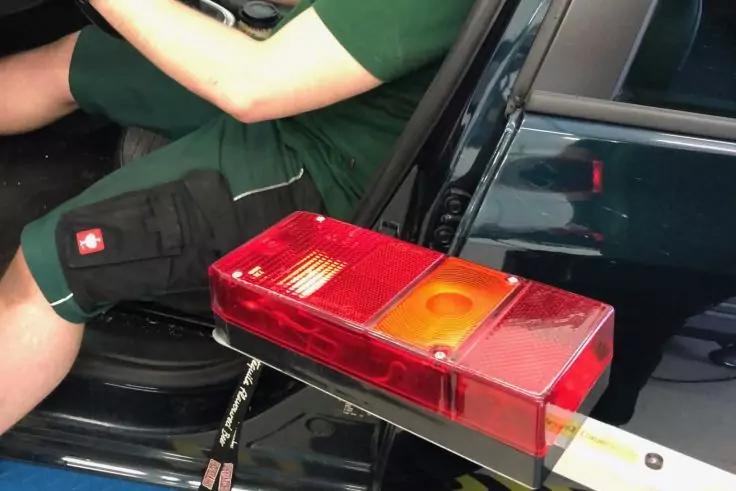 Rote und orange rechteckige Kunststofflinse auf weißer Stange neben einer offenen Autotür; am Bildrand der Unterkörper einer sitzenden Person in grünem Oberteil und schwarzen Shorts mit rotem Quadrat und weißem Kreuz, Schlüsselbund sichtbar