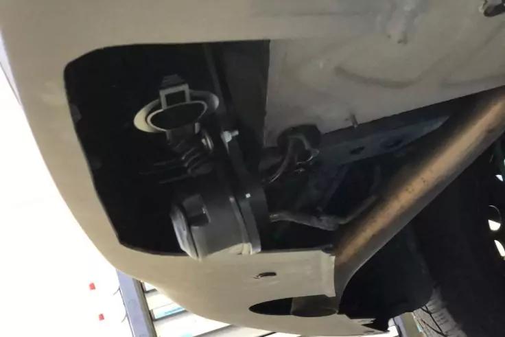 Unterseite eines Fahrzeugs mit Metallrahmen, Metallrohr, schwarzer Kunststoffschürze, Halterung mit runder Abdeckung und teilweise sichtbarem Reifen