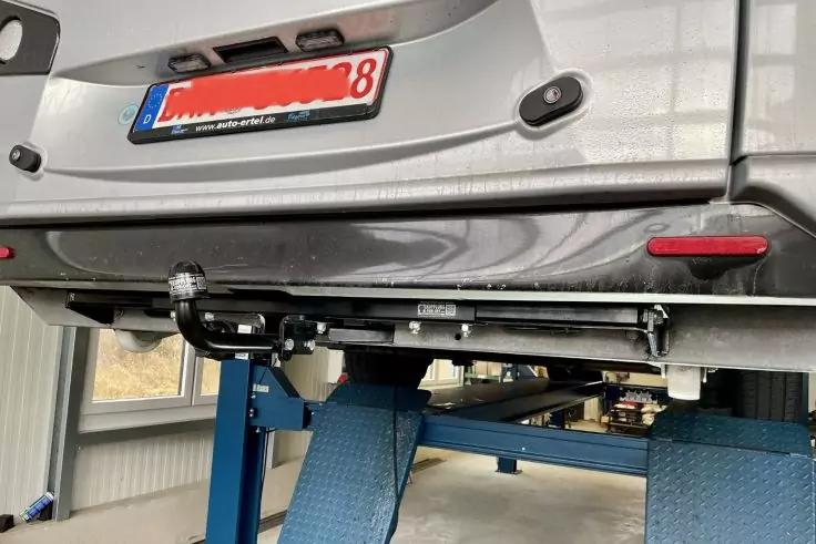 Heck eines silbernen Autos auf einer Hebebühne mit sichtbarer Anhängerkupplung, teilweise unkenntlichem Kennzeichen und blauen Hebebühnenarmen in einer Werkstatt