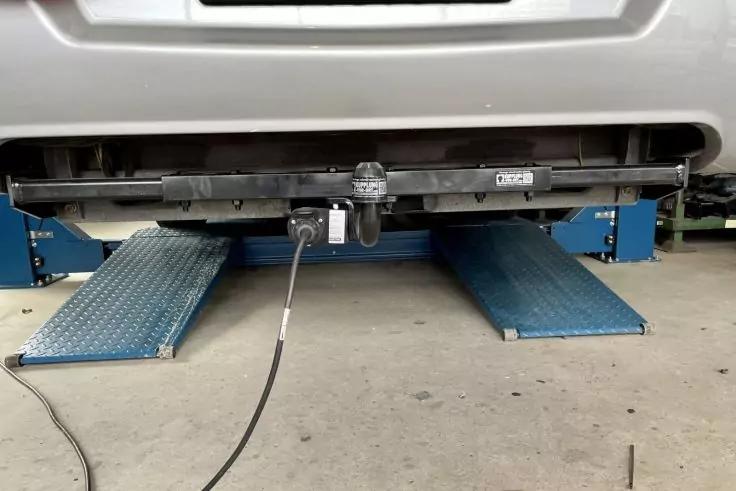 Heck eines Fahrzeugs mit Stoßstange und Anhängerkupplung, an der ein Stecker mit Kabel angeschlossen ist; darunter Metallrampen einer Hebebühne und Betonboden