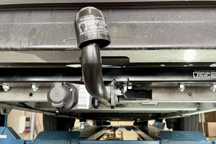 Unterseite des Heckbereichs eines Fahrzeugs auf einer Hebebühne, sichtbare Anhängerkupplung mit Schutzkappe, Anhängersteckdose, Fahrzeugchassis und Teile der Hebebühne