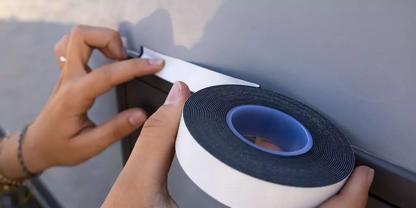 Zwei H&auml;nde bringen eine Rolle wei&szlig;es Schaumstoffklebeband mit schwarzer Schaumstoffschicht auf einer glatten grauen Oberfl&auml;che an