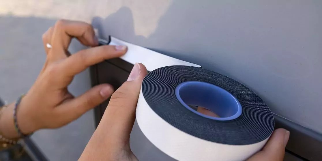 Zwei H&auml;nde bringen eine Rolle wei&szlig;es Schaumstoffklebeband mit schwarzer Schaumstoffschicht auf einer glatten grauen Oberfl&auml;che an